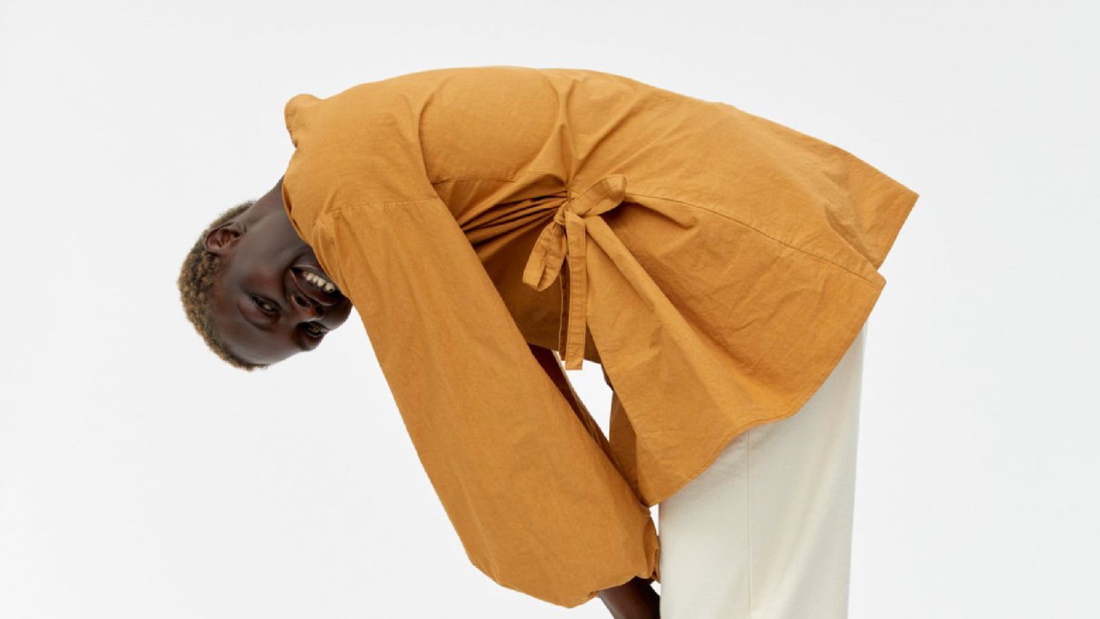 Portrait éditorial en studio : un mannequin rit, penché en avant, dans un ensemble ample ocre et un pantalon écru — image représentative de la créativité de la mode nordique.